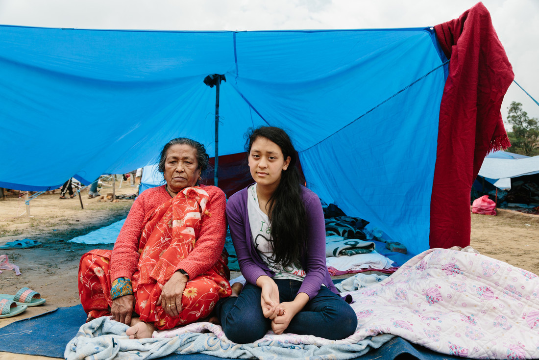 An elderly woman and a girl sit on blankets in front of a tarpaulin.