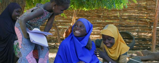Children smiling at each other while sitting outside in the shadow with their schoolbooks.