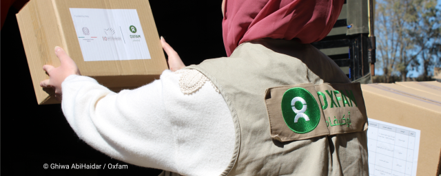 A person wearing an Oxfam vest receives a cardboard aid box from someone standing in a truck during a humanitarian supply distribution. Trees and blue sky are visible in the background.
