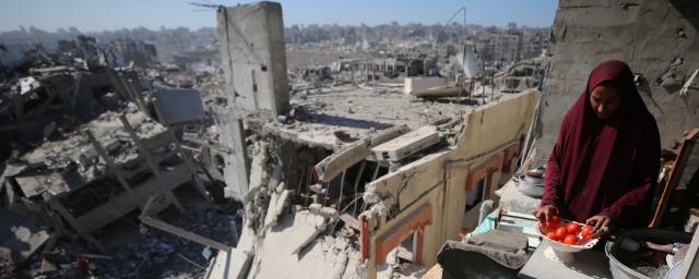 A woman sorting tomatoes on a damaged structure in Gaza, with destroyed buildings in the background.