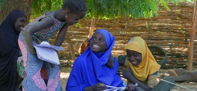 Children smiling at each other while sitting outside in the shadow with their schoolbooks.