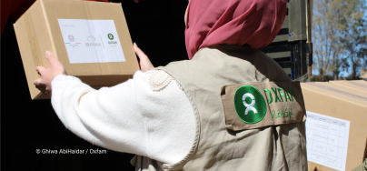 A person wearing an Oxfam vest receives a cardboard aid box from someone standing in a truck during a humanitarian supply distribution. Trees and blue sky are visible in the background.