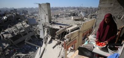 A woman sorting tomatoes on a damaged structure in Gaza, with destroyed buildings in the background.